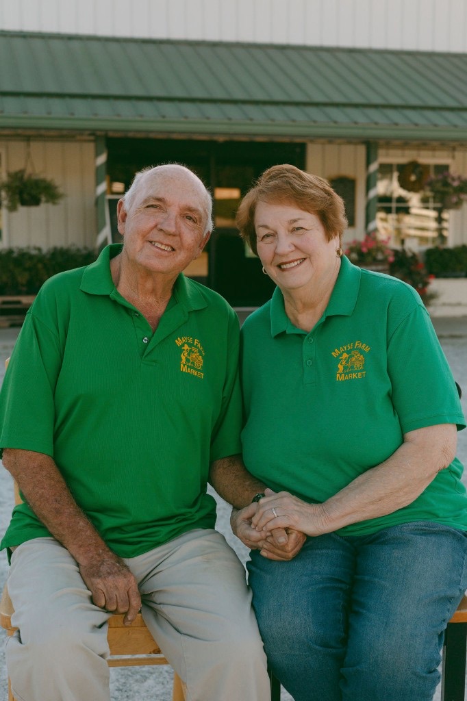 Paul and Sherrill Mayse sitting together in matching Mayse Farm Market shirts in front of the market