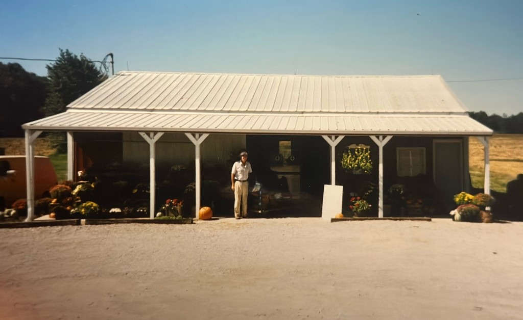 The farm market with its wide front porch, mums and pumpkins out front, and a man standing under the roof